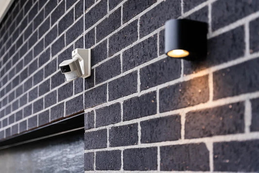 A PSA Centrii Sensor Camera mounted on a dark brick wall next to a modern outdoor wall light